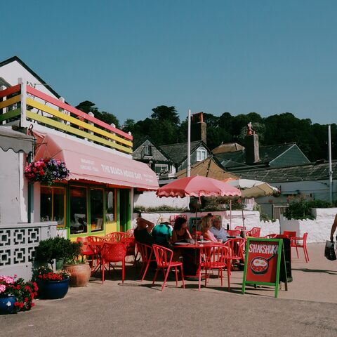 The Beach House Cafe in Lyme Regis with pink awning, colourful railings, red umbrellas, and a Shakshuka menu board out front.