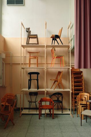 A tall pine rack displays eight chairs one per shelf, with more chairs stacked on the floor of a hall with peach walls and a mauve curtain.