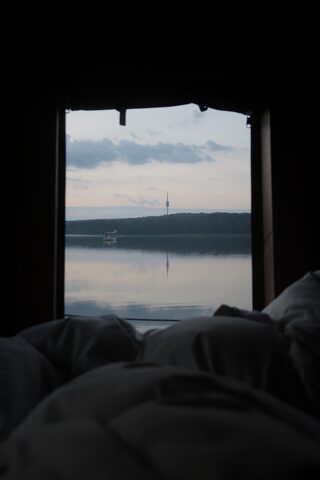 Dawn view from bed on a raft, through the open cabin end across still water to a wooded shore, with the Fernmeldeturm Berlin-Schäferberg on the horizon.