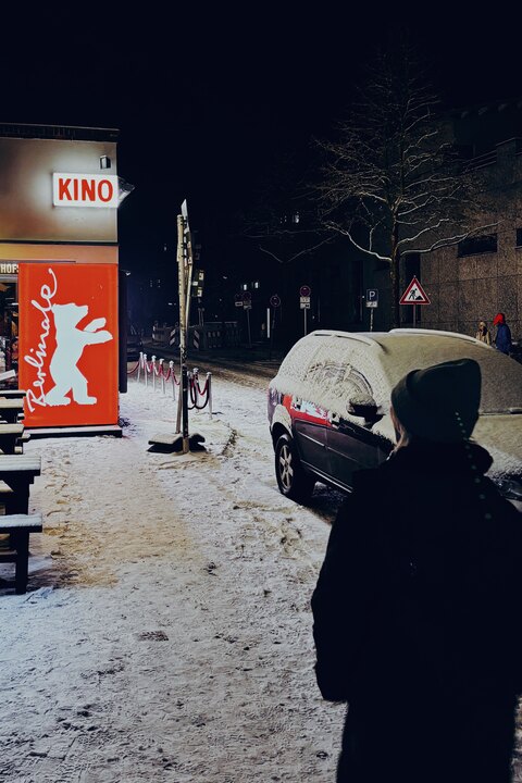 Snowy night street scene in Berlin beside a red Berlinale sign, with a snow-covered parked car and a person in the foreground.
