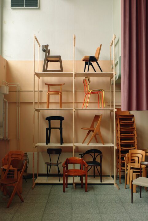 A tall pine rack displays eight chairs one per shelf, with more chairs stacked on the floor of a hall with peach walls and a mauve curtain.