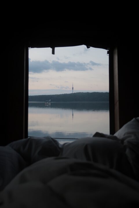 Dawn view from bed on a raft, through the open cabin end across still water to a wooded shore, with the Fernmeldeturm Berlin-Schäferberg on the horizon.