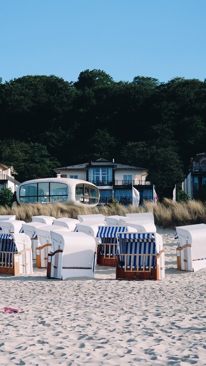 The white, shell-shaped Müther-Turm — the former lifeguard tower on the Südstrand beach in Binz on Rügen — rising behind rows of striped Strandkörbe beach chairs on the sand, with a forested hillside and clear blue sky beyond.