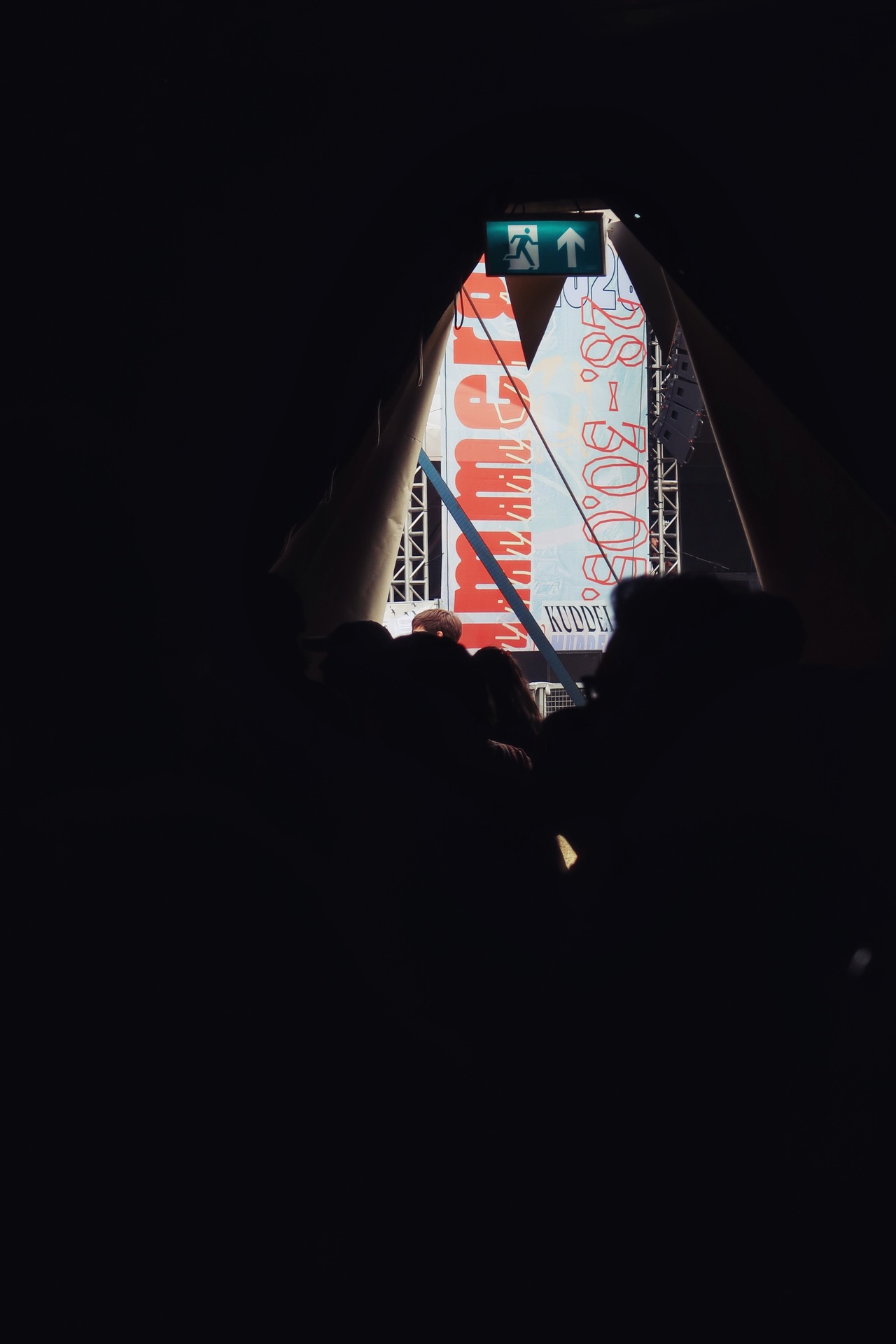 View from inside the dark stage tent, looking out through the triangular opening at the Immergut Festival 2026 main-stage banner in the bright daylight outside, with silhouetted festival-goers in the foreground and a green emergency exit sign above.