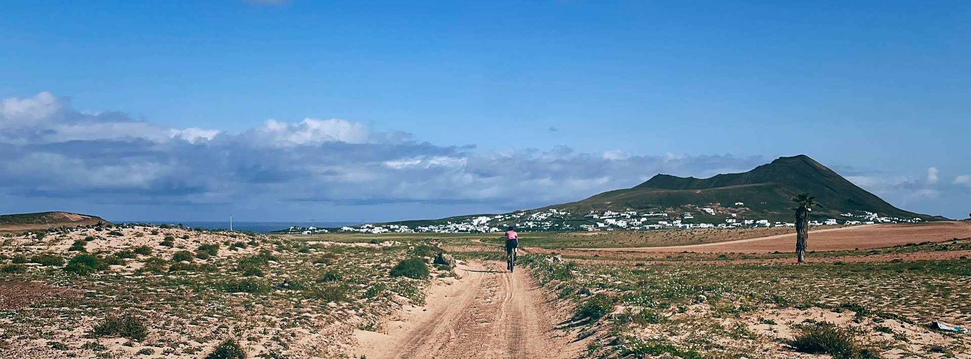 A cyclist rides a dirt track through dry fields toward the village of Soo and nearby volcanic hills beneath a wide blue sky.